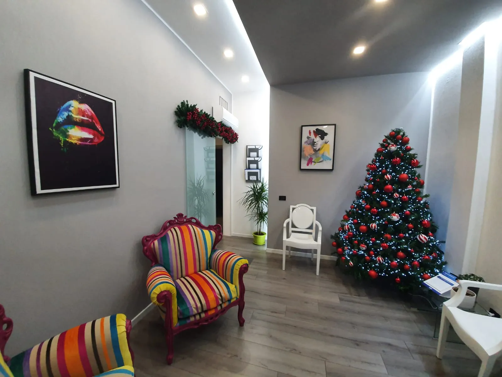 La sala d'attesa: poltrone barocche a righe colorate, un quadro pop art con due labbra, sedie bianche e l'albero di Natale accanto alla porta in vetro.
