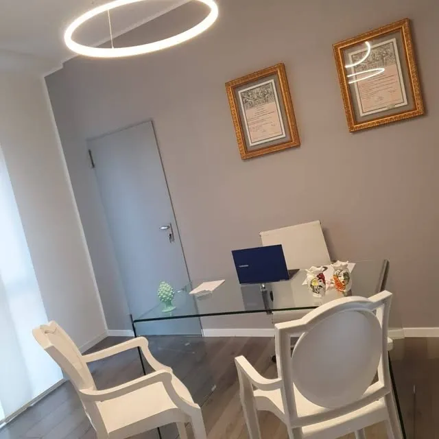 L'ufficio, con la scrivania in vetro, le sedie bianche, una lampada ad anello e due diplomi alle pareti.